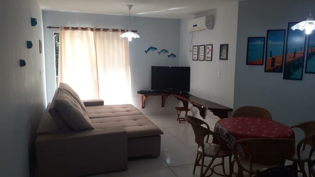 Sala de estar do Lindo Apto Praia da Tabatinga com mesa redonda de jantar do lado direito da imagem com quatro cadeiras, um sofá do lado esquerdo e em frente ao sofá uma cômoda e ao lado esquerdo da cômoda uma TV.