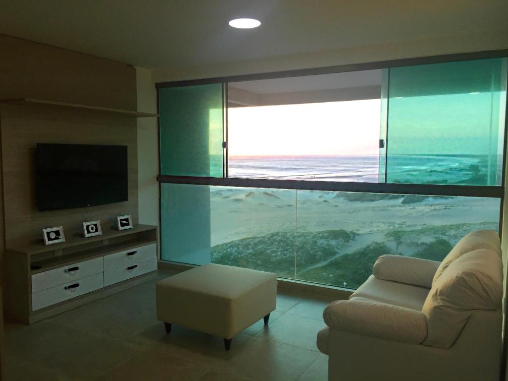 Sala de estar do airbnb Orla Praia Grande. No canto direito da sala há um sofá, no meio há um puff e no canto esquerdo há um rack com uma TV presa. No fundo da sala há uma parede feita de vidro que possui uma janela grande com vista para a praia que está bem próxima.