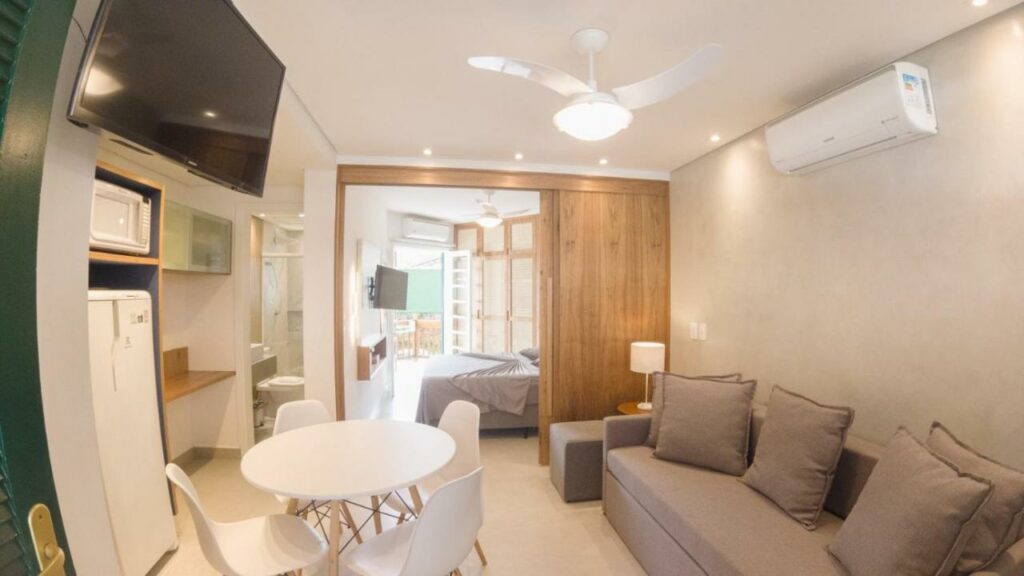 Imagem do ambiente do Porto Paúba Flat 116 com sala de estar a frente com sofá do lado direito da imagem, em frente ao sofá uma mesa redonda com quatro cadeiras e  do lado direito da mesa uma cozinha compacta. Representa airbnb na praia de Santiago.