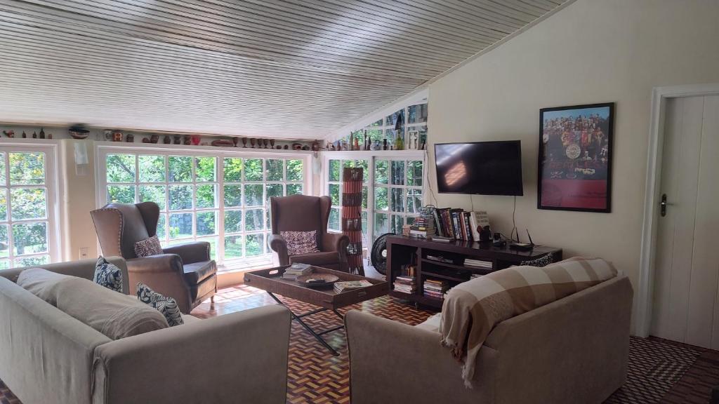 Sala de estar do airbnb Rancho São Carlos. No canto direito é possível ver um pedaço de uma porta e no lado esquerdo há um rack com livros e uma TV presa a parede. No lado esquerdo da sala a parede é composta por três janelas bem grandes. Ainda no canto esquerdo há duas poltronas grandes. No centro da sala há uma mesa de centro e no lado direito há dois sofás.