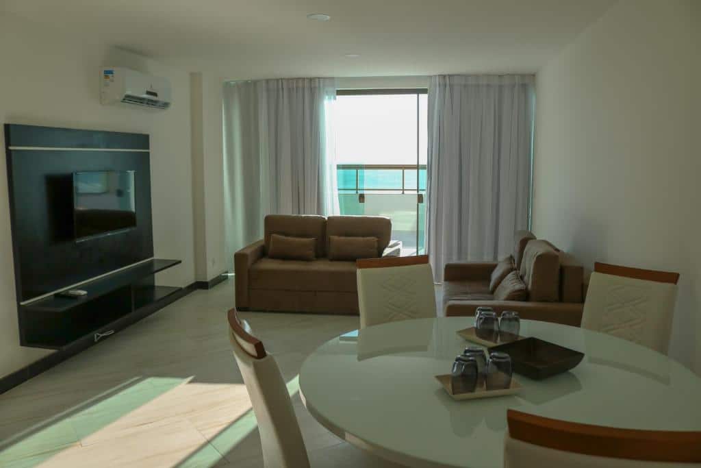 Sala de estar do airbnb Sophia II Residence Inn. No canto direito da imagem há uma mesa de jantar redonda com cadeiras em volta, atrás da mesa há dois sofás e na parede do lado esquerdo há um rack com uma TV presa nele. No fundo da sala há uma grande porta de vidro que dá acesso a varanda e oferece vista para o mar.