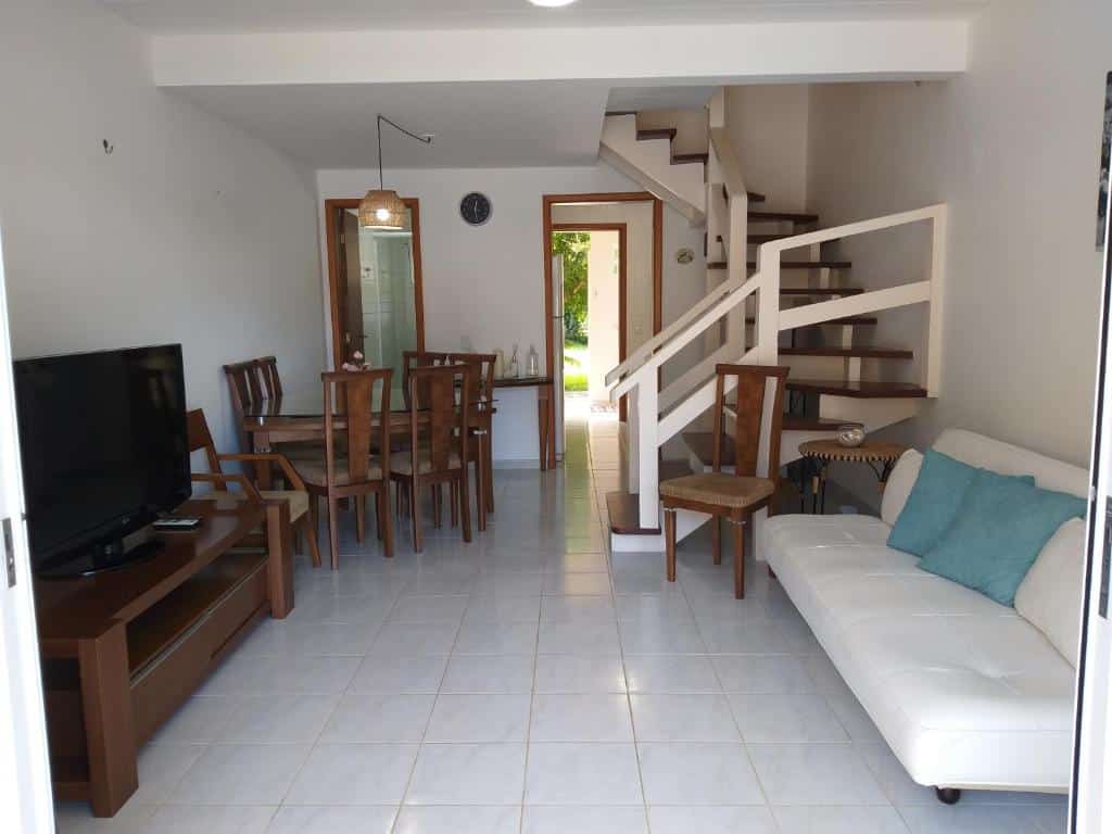 Sala de estar do Villagio Charmoso na Praia da Mococa com sofá do lado direito em frente ao sofá do lado esquerdo uma cômoda com TV, ao fundo do lado esquerdo da cômoda com a TV uma mesa de madeiras com seis cadeiras.