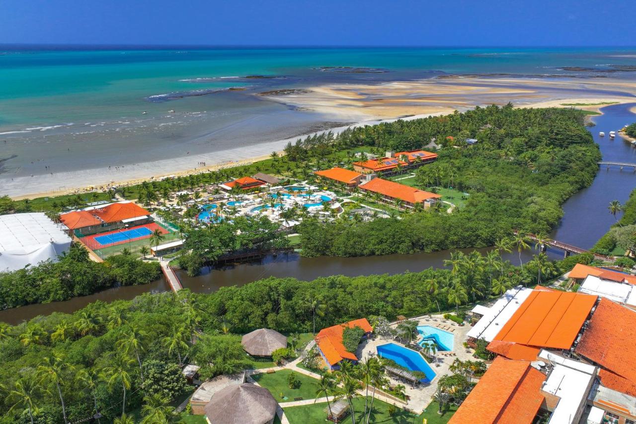 A imagem mostra a vista aérea do Salinas Resort, em Maragogi, luxuoso, localizado entre uma praia de águas azul-turquesa e um rio cercado por vegetação densa. O resort é composto por vários edifícios com telhados vermelhos e laranja, dispersos entre áreas verdes bem cuidadas. Na parte voltada para a praia, há uma grande piscina azul próxima de uma série de palmeiras, enquanto outra piscina é visível mais ao centro, rodeada por um complexo de toboáguas coloridos e áreas de lazer. No centro da imagem, há pontes pequenas sobre o rio que permitem a circulação entre diferentes partes do resort.