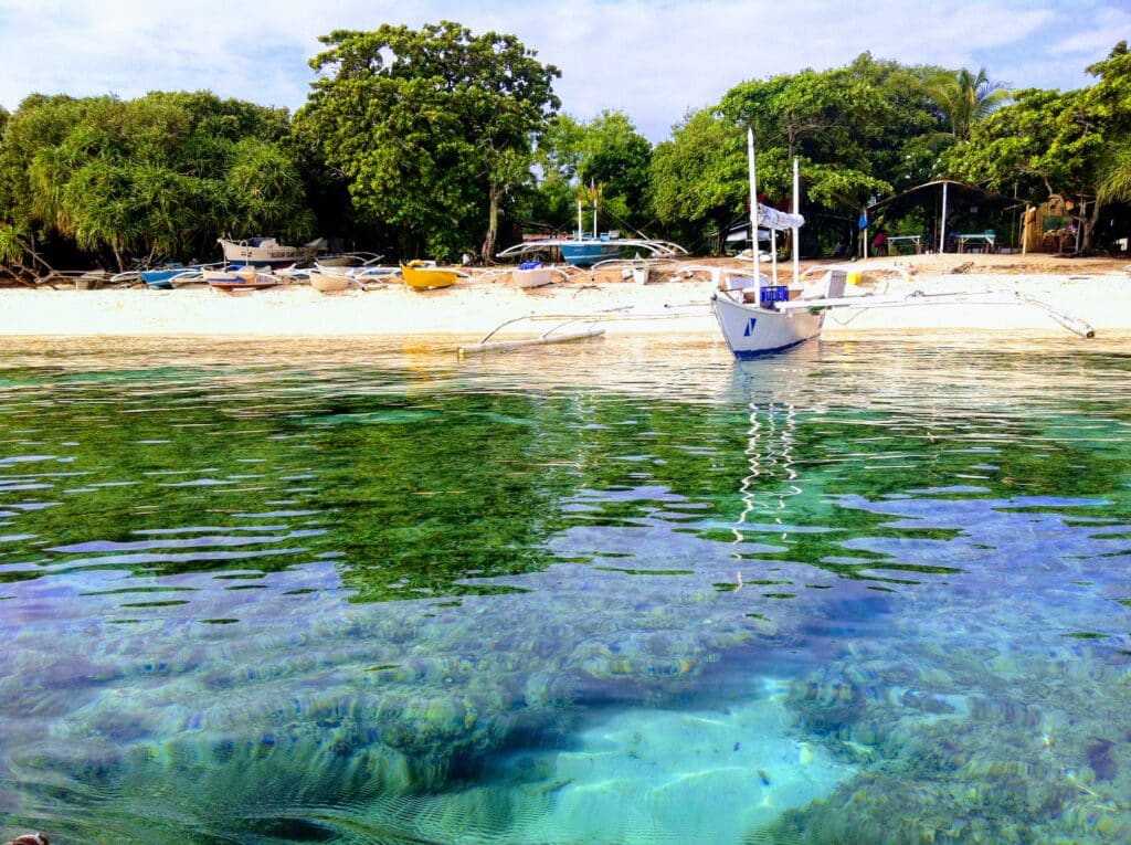 Um pequeno barco de madeira flutua em águas cristalinas azul-turquesa perto da praia de Balicasag, arenosa cercada por árvores verdes e vários barcos atracados. Representa o post sobre melhores praias nas Filipinas.