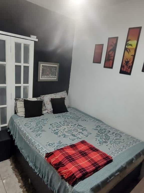 Foto do quarto em Serra Negra - Melhor localização da cidade. Há uma cama de casal com manta e travesseiros. Na parede a qual se encosta à direita há pequenos quadros, e atrás há uma janela.
Ilustra o post sobre airbnb em Serra Negra.