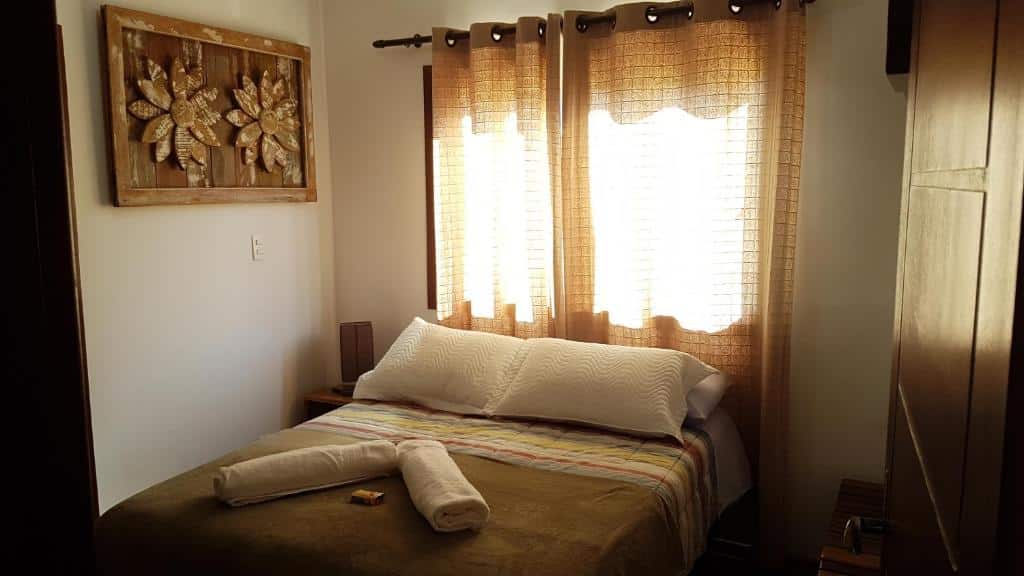 Foto do quarto do Solar Douro. Ilustra o post sobre airbnb em Ouro Preto. A cama está no centro, e atrás dela há uma janela com cortinas. Do lado direito da cama há uma cama e um quadro decorando a parede. Já na esquerda há um guarda-roupa.