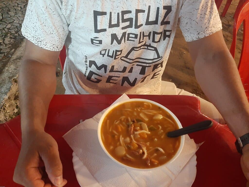 Homem sentado, aparecendo na imagem do seu ombro para baixo. Ele está usando uma camiseta escrito "cuscuz é melhor que muita gente" e com os braços apoiados em uma mesa que tem um isopor redondo com sopa dentro