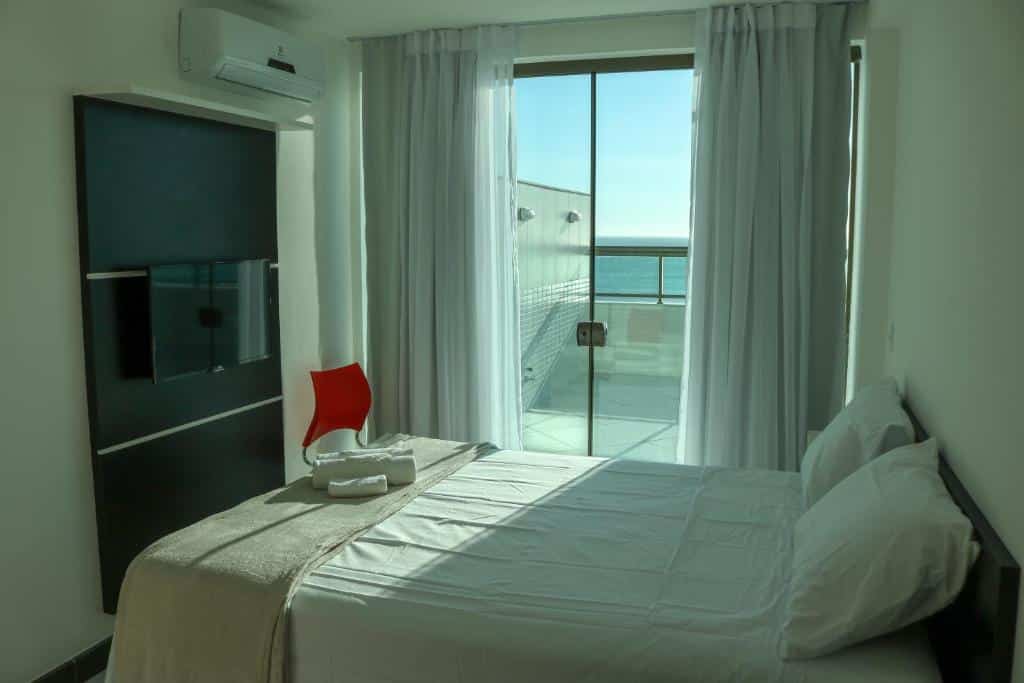 Quarto do airbnb Sophia II Residence Inn. A cama de casal está encostada na parede do lado direito da imagem e logo a frente da cama há uma TV pendurada na parede. No canto esquerdo do quarto há uma cadeira e ao fundo uma grande porta de vidro que dá acesso para a varanda e possui vista para o mar. Imagem utilizada para ilustrar o post airbnb em Arraial do Cabo.