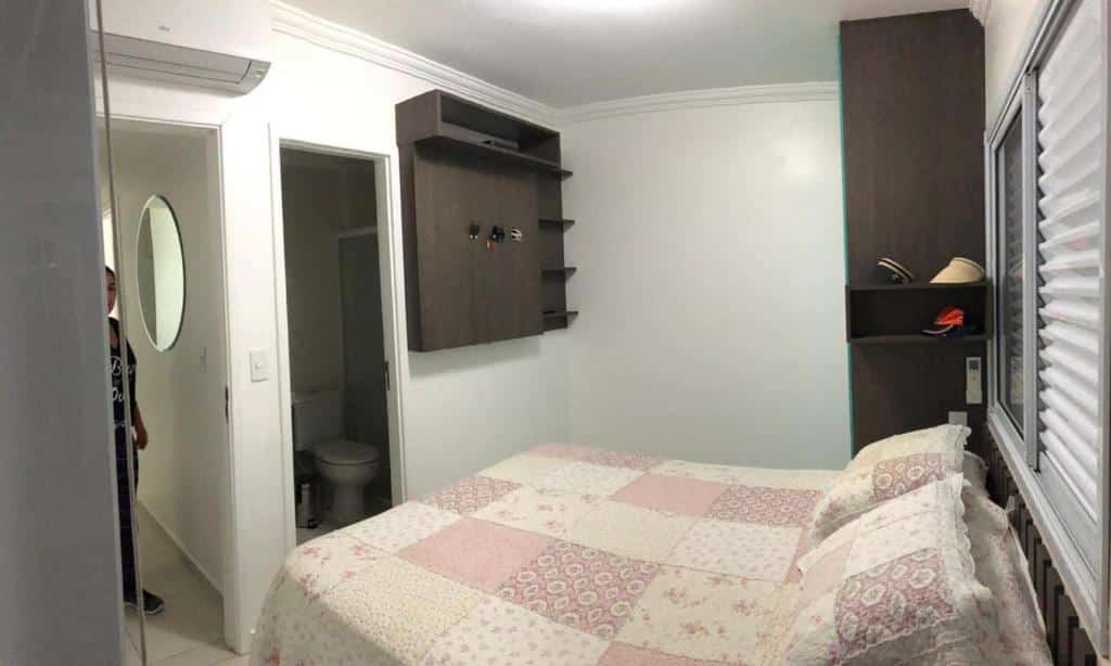 Quarto do Spazio Felicita, um dos airbnb em Bertioga. Uma cama de casal com colcha e travesseiros está embaixo de uma janela na parede direita. Ao lado direito fica um armário, e em frente está a porta que leva ao banheiro e o aparelho de ar-condicionado.