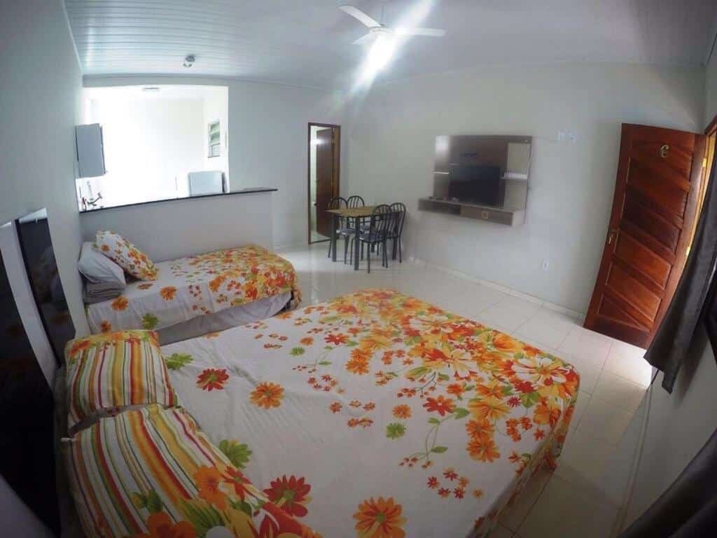 Quarto e sala do airbnb Suítes do Cid. no canto direito está a porta que dá acesso a saída do airbnb, no meio do cômodo há uma cama de casal no lado direito e uma cama de solteiro no lado esquerdo, na frente das camas há uma TV e uma mesa com cadeiras. No lado esquerdo há um balcão que divide o cômodo da cozinha. Imagem para ilustrar o post airbnb em Ilha Grande.