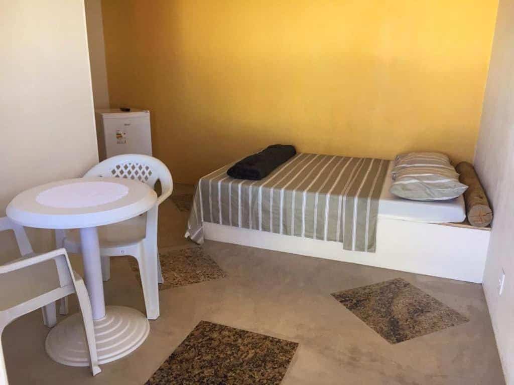 Quarto do Suítes Pé Nareia Itaipuaçu. Uma cama do lado direito, do lado esquerdo uma mesa de plástico com duas cadeiras e um frigobar no fundo.