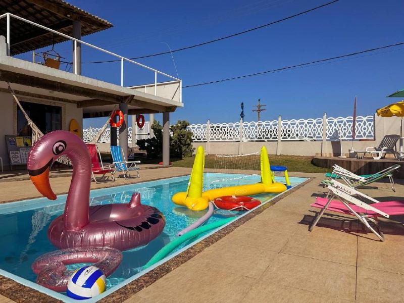 Área externa do Suítes Pé Nareia Itaipuaçu. Uma piscina no meio com boias e brinquedos infantis, ao redor cadeiras de praia.