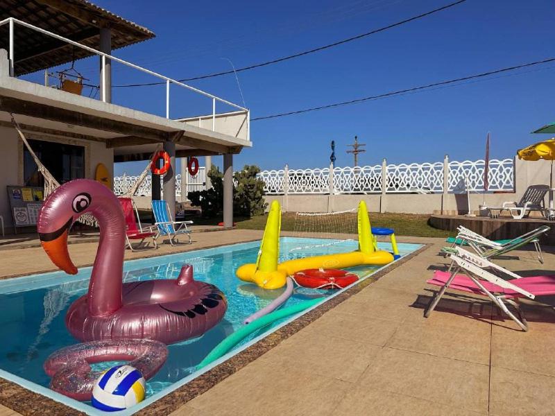 Área externa do Suítes Pé Nareia Itaipuaçu. Uma piscina no meio com boias e brinquedos infantis, ao redor cadeiras de praia.