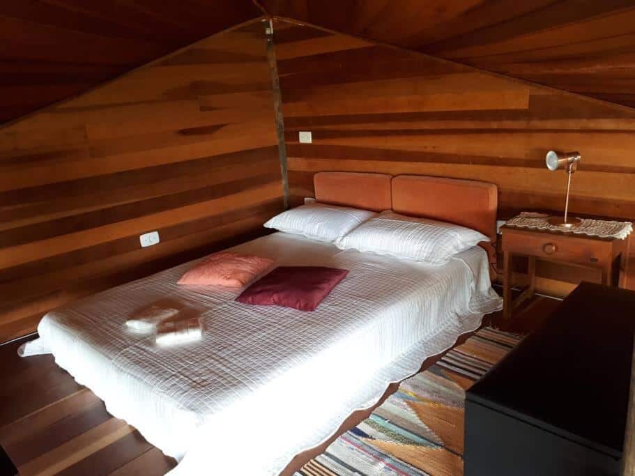 Foto do quarto do Tao da Serra – cabana em meio à natureza!. Representa o post sobre airbnb em São Francisco Xavier. A cama de casal está com travesseiros e almofadas em cima, e do lado direito dela há uma mesa e cabeceira com abajur em cima.
