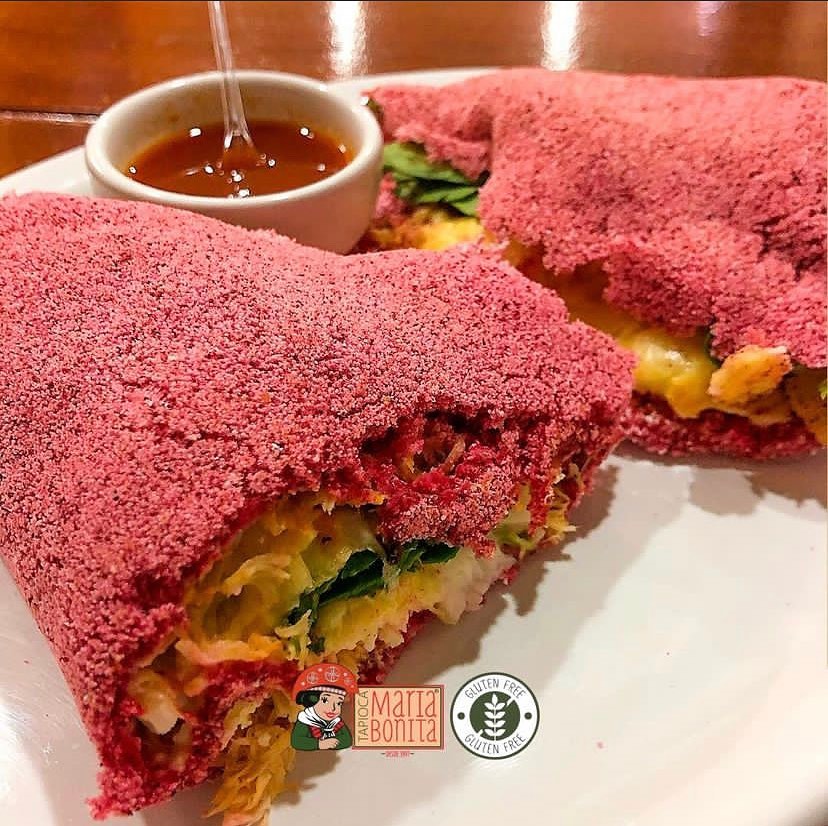 Tapioca com massa rosa feita de beterraba em um prato branco com um potinho com molho de pitanga com laranja. Representa um dos restaurantes em Maceió