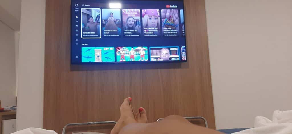 Pernas de uma mulher com esmalte vermelho cruzada em cima da cama, com uma TV ligada no You Tube na frente.