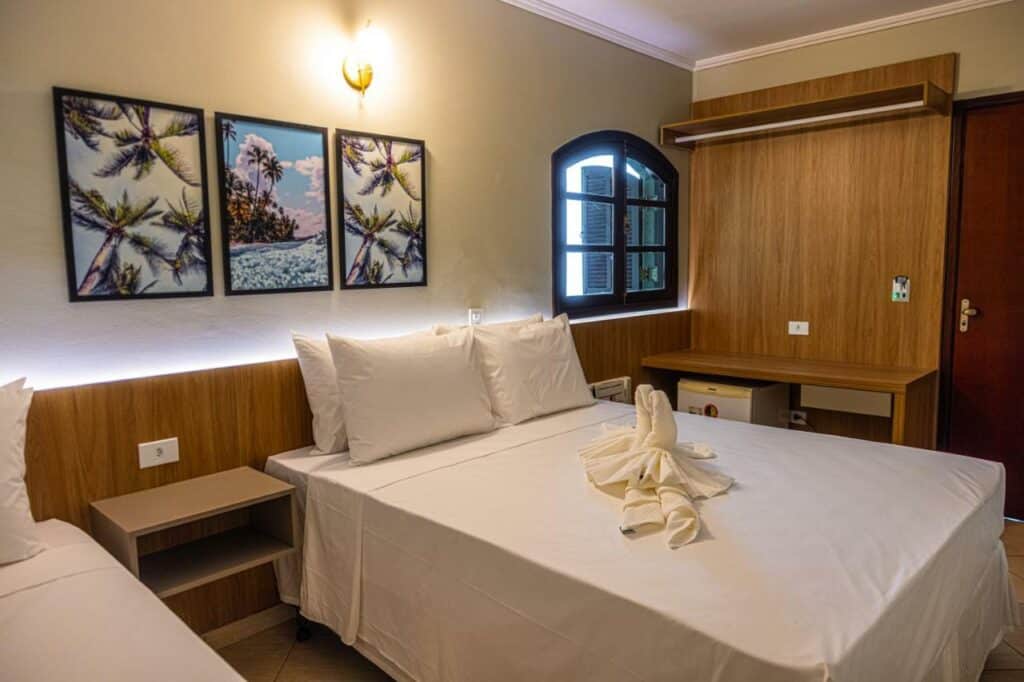 Quarto triplo no Ubatuba Eco Hotel, com uma cama de casal, uma cama de solteiro, três quadros decorativos com imagens de litoral, e toalhas em formato de cisne sobre a cama