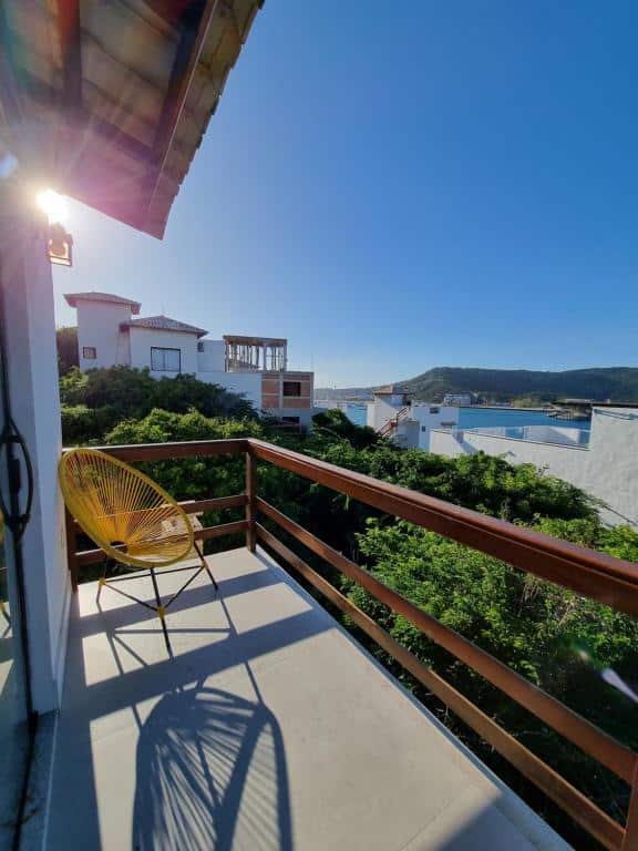 Varanda do airbnb Casa Corazul. A varanda é pequena e o parapeito é feito de ripas de madeira. No canto esquerdo da varanda há uma cadeira. A varanda oferece vista para algumas árvores, algumas casas com muros brancos e ao fundo é possível ver o mar.