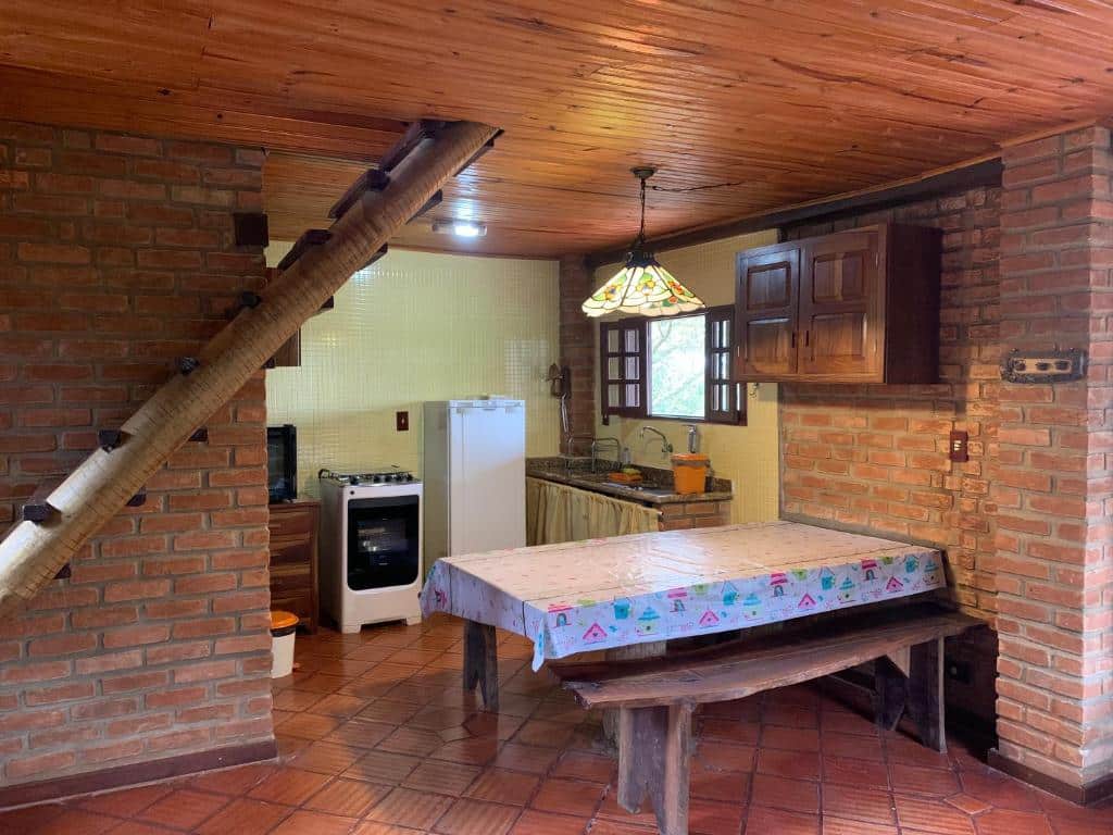 Parte do Varandas da Serra / Reserva Alto da Colina que mostra do lado direito a cozinha do chalé com uma mesa retangular com um banco de madeira, um armáerio na parede de tijolos ao lado direito da mesa, e ao fundo uma pia pra lavar louças, uma janela aberta, uma geladeira e do lado um fogão. Já na parte esquerda da foto tem um tronco de madeira com degraus para subir no segundo andar. Imagem ilustrando post Airbnb em Ibitipoca.