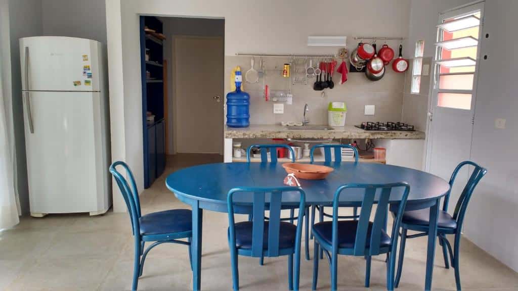 Cozinha da Vila Juma com uma longa mesa de jantar com seis cadeiras no centro do cõmodo. Ao fundo está a bancada da pia com fogão, utensílios e um galão d'água. Na lateral esquerda há uma geladeira.