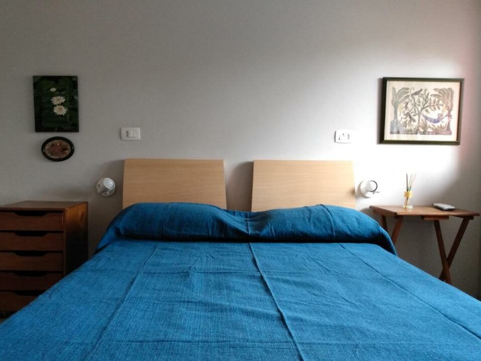 Quarto da Vila Juma, um dos airbnb em Camburizinho. Uma cama de casal tem mesinhas de cabeceira, luminárias e quadrinhos dos dois lados.