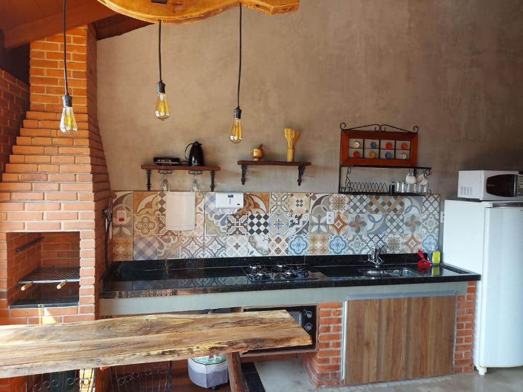 Parte do Villa da Serra chalé que mostra a cozinha com uma mesa de madeira, e atrás uma churrasqueira, do lado um cooktop e um forno embaixo. Há também uma pia de mármore preto ao lado do cooktop, uma geladeira com um micro-ondas em cima e algumas prateleiras na parede. Imagem ilustrando post Airbnb em Ibitipoca.