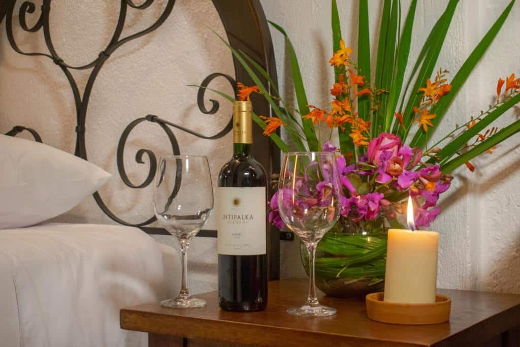 garrafa de vinho com duas taças de vidro em cada lado, além de um arranjo de flores e uma vela em cima de uma pequena cômoda de madeira, ao lado de uma cama de casal no vila insight hotel barato machupicchu