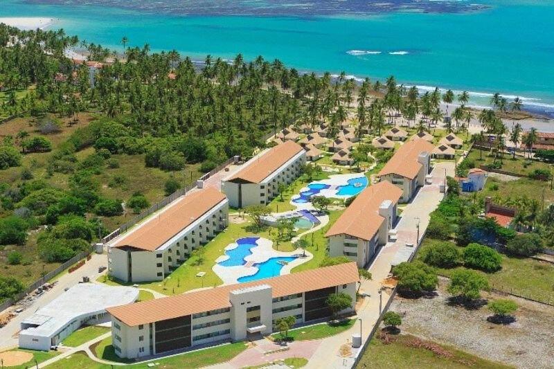 A imagem mostra uma vista aérea do Carneiros Beach Resort, localizado à beira-mar. O resort é composto por vários edifícios brancos longos distribuídos de maneira ordenada ao longo do complexo, todos com telhados de cor clara. No centro do resort, várias piscinas de formatos sinuosos e azuis estão interligadas, cercadas por numerosas espreguiçadeiras e guarda-sóis. Palmáceas e uma vegetação tropical densa circundam os edifícios e piscinas, adicionando um toque natural ao ambiente. A praia de areia clara e as águas azul-turquesa do oceano estendem-se ao longo do lado esquerdo da imagem, contrastando com o verde exuberante das palmeiras e arbustos que isolam o resort do ambiente externo. Representa resorts em Maragogi