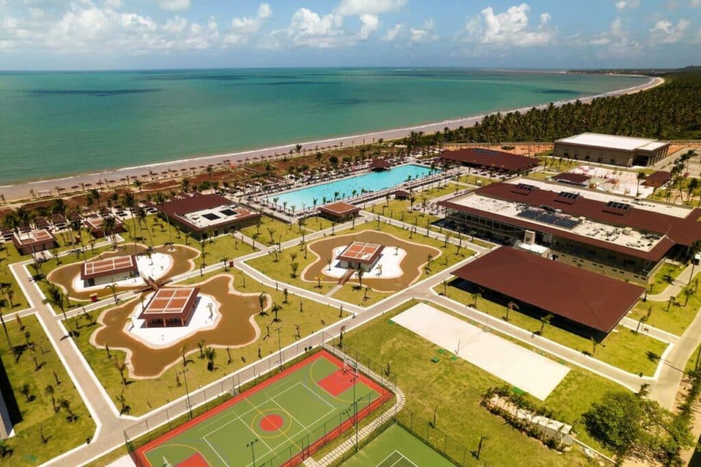 Vista aérea do Vila Galé Resort Alagoas. O resort tem várias áreas, incluindo uma piscina extensa, quadra de esportes e telhados mostrando os prédios do local. Há bastantes gramados nos arredores dos prédios, árvores e o resort fica à beira-mar