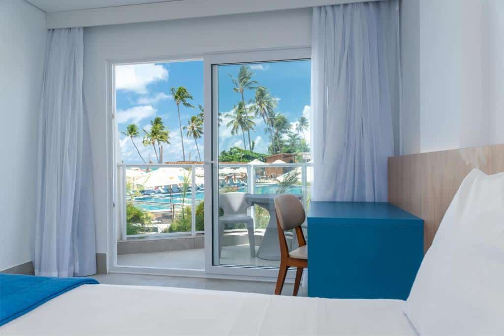 Quarto Premium Duplo do Maragogi Brisa Exclusive Hotel, com uma mesa de trabalho no interior e mostrando a janela de vidro com as cortinas abertas que dá acesso à sacada com vista da piscina e coqueiros
