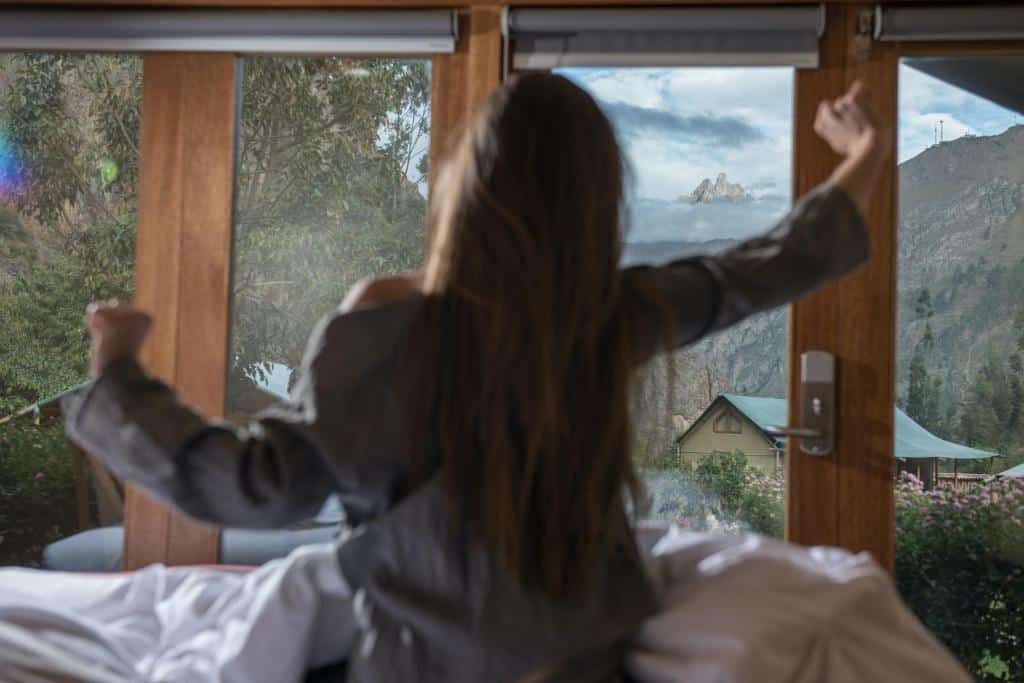 mulher de costas para a câmera se espreguiçando em cima da cama, olhando para as paredes de vidro do quarto que dão vista para as montanhas peruanas, na região do Valle Sagrado, no Peru.