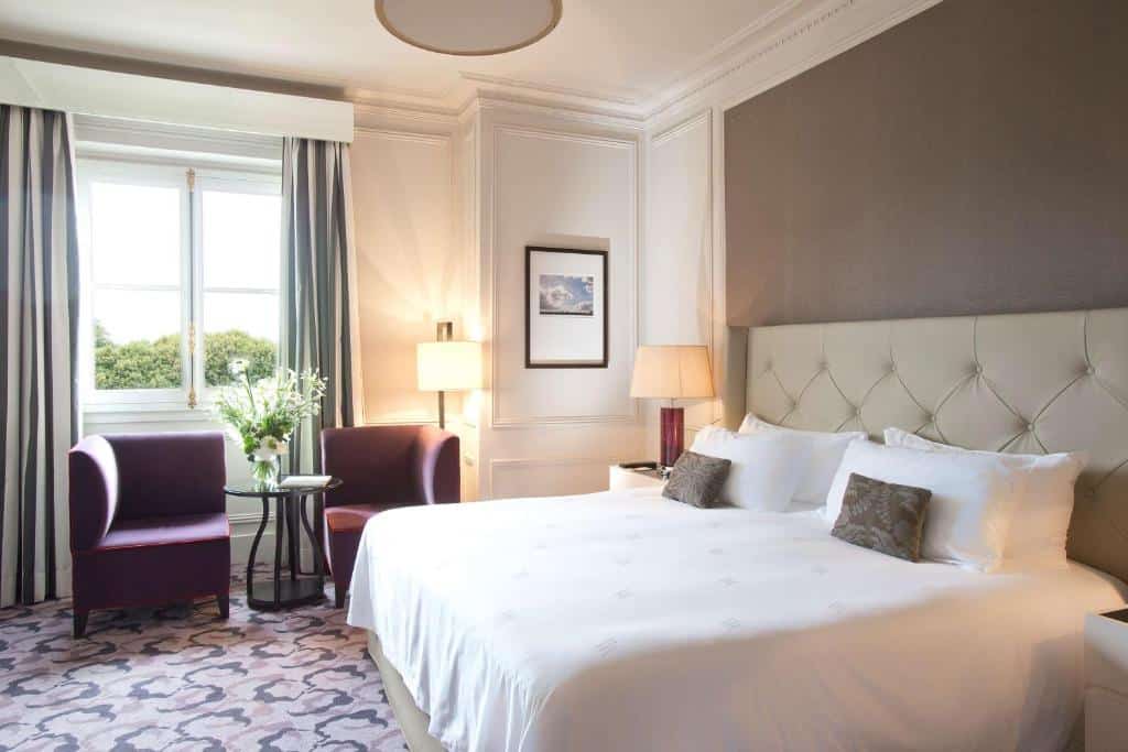 Quarto do Waldorf Astoria Versailles, um dos hotéis perto do Palácio de Versalhes. Uma cama de casal está encostada na parede ao lado direito e possui mesinhas de cabeceira e abajures dos dois lados. Na parede ao lado da cama está uma janela com vista do jardim, e do lado de dentro fica uma área de estar com poltronas, mesinha, abajur e um vaso de flores. Há um quadrinho na parede ao lado da janela.