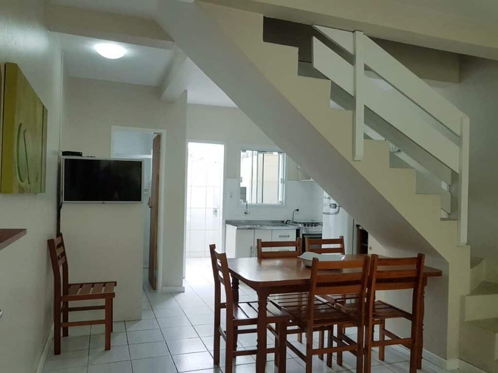Visão geral do RESIDENCIAL COM CASAS DE 2 DORM - Praia Garopaba. Uma mesa de jantar com seis cadeiras fica logo abaixo da escadaria, e ao fundo está a cozinha.