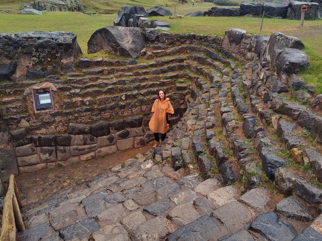 mulher usando uma capa de chuva laranja no centro da imagem, ao seu redor há vários degraus de pedras de granito no sítio arqueológico de Saqsayhuaman, em cusco.