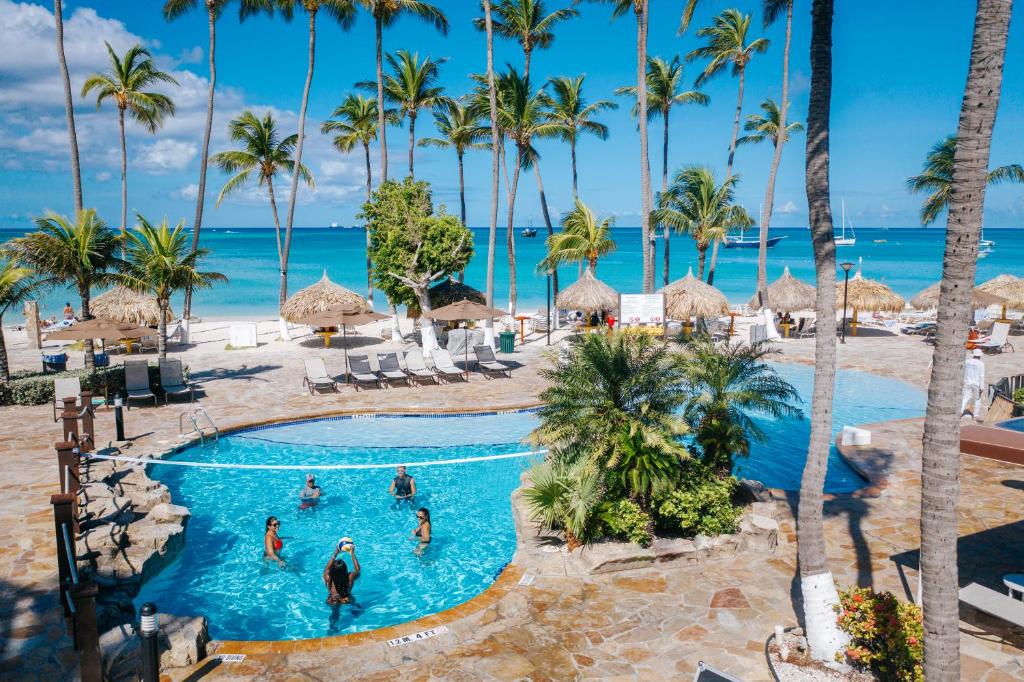 Piscina externa do All Inclusive Holiday Inn Resort Aruba. Ela está no meio de uma área aberta, rodeada de váras espreguiçadeiras, guarda-sóis e algumas árvores. Logo em frente está o mar.