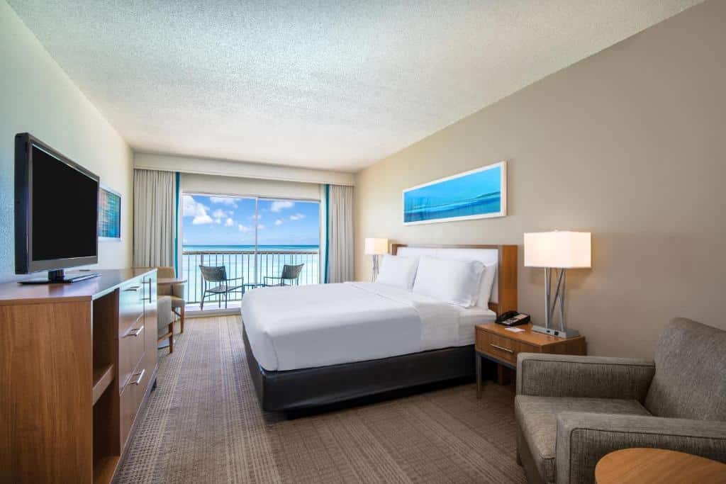 Quarto do All Inclusive Holiday Inn Resort Aruba. Uma cômoda, uma televisão, duas poltronas e uma mesinha do lado esquerdo. Do lado direito uma poltrona, uma cama de casal e um abajur com uma cômoda de cada lado. No fundo a varanda com vista para o mar.