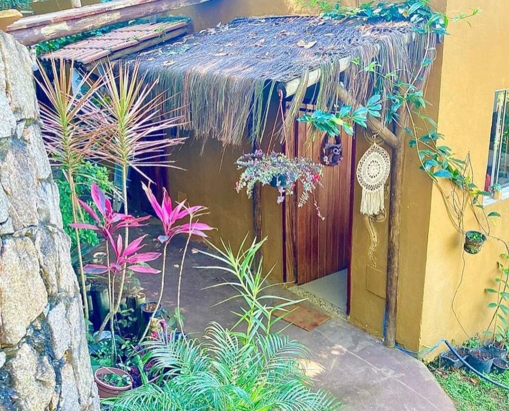 Entrada do Altar da praia Vermelha 180° de vista para o mar. Ela está no térreo, é uma porta de madeira e umm pequeno telhado de palha, Flores e plantam estão rodeando o local.