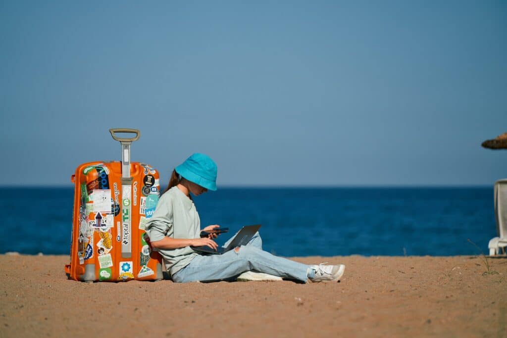 Uma mulher sentada na areia de uma praia mexendo em um notebook junto de uma mala de viagem, para representar os viajantes nômades da SafetyWing