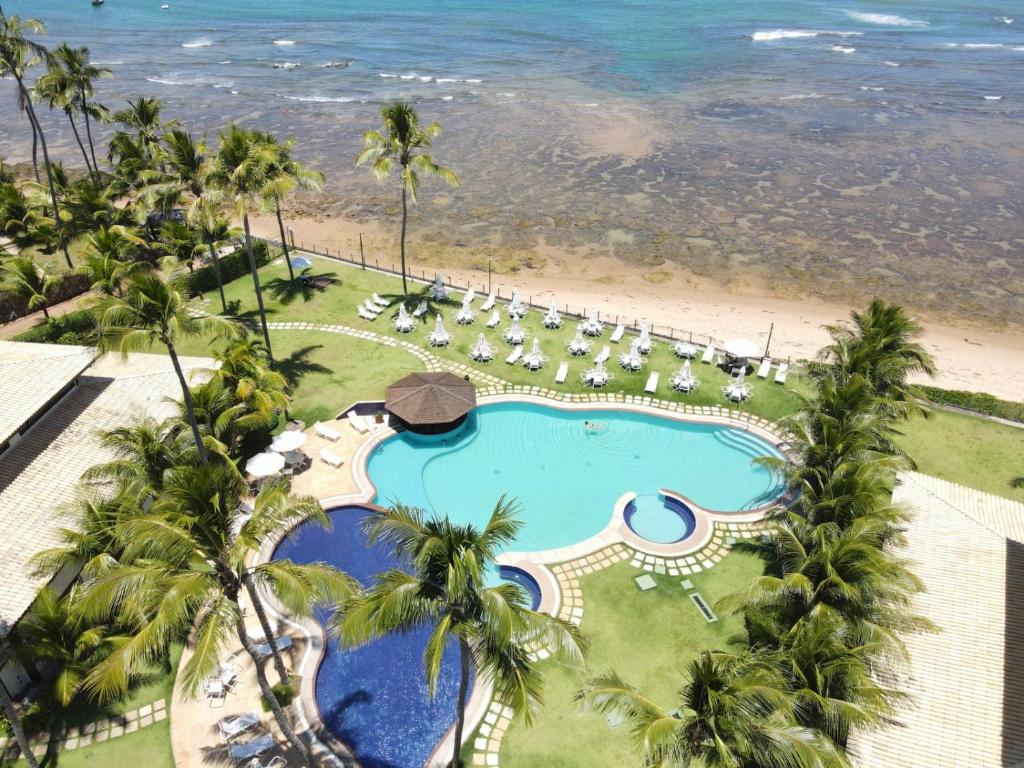 Vista aérea do Praia do Forte - ap Bali Bahia 107. Há duas piscinas numa grama vem verde, quiosque e diversas espreguiçadeiras. Há muitas palmeiras e coqueiros, e a praia está logo à frente.