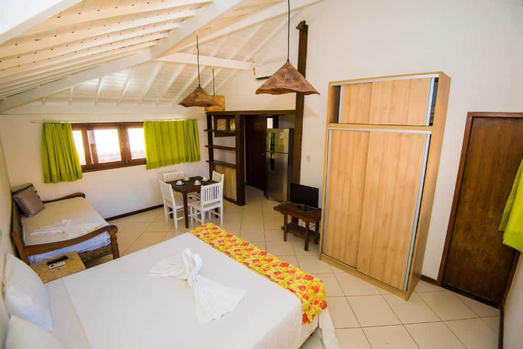Foto do quarto em Apart Hotel Forte Blu, que ilustra o post de airbnb na Praia do Forte. Há uma cama na esquerda, e outra cama de solteiro ao seu lado. N frente da cama há um guarda-roupa e uma tv. No fundo há uma mesa pequena com quatro lugar e uma cozinha compacta.