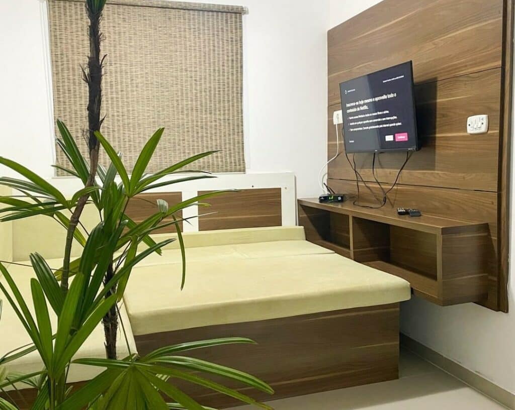Sala de uma das acomodações dos Apartamentos da 23. A televisãp está pendurada em um painel do lado direito. Do lado esquerdo e no meio da imagem está o sofá. Uma planta está no canto da imagem. Ele também faz parte da lista de airbnb na Praia do Sapê.