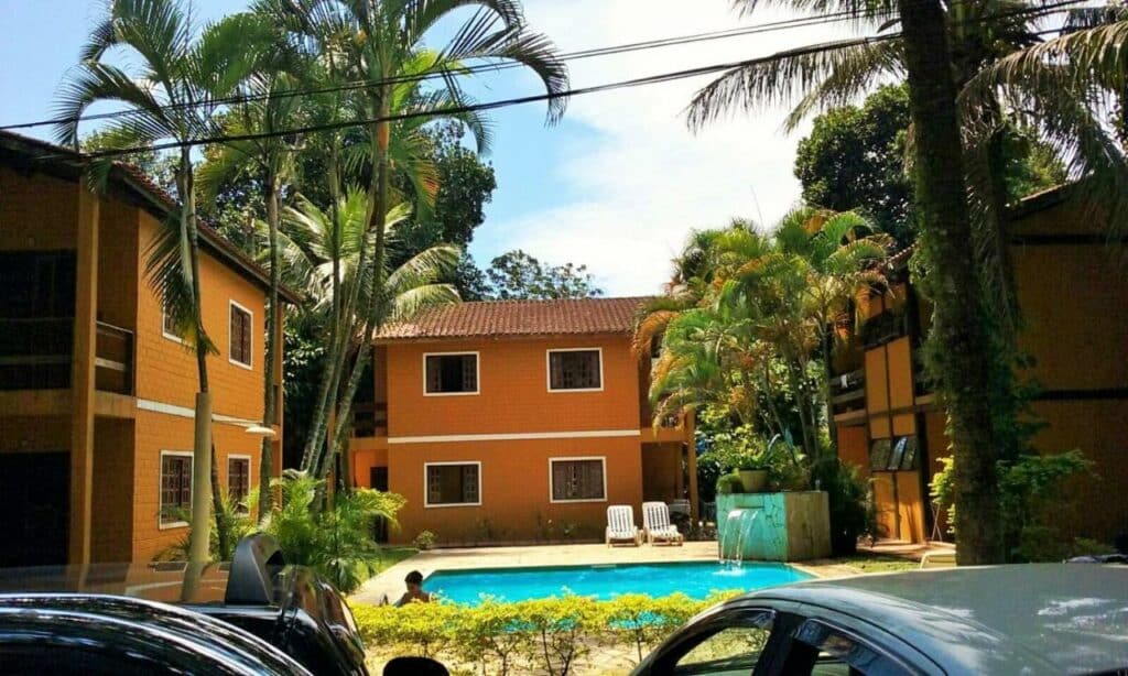 Lado de fora do Apartamento Condomínio Boiçucanga Flat N 39 - Apto 10. Vários apartamentoos estão ao redor de uma piscina. No canto da imagem é possível ver também alguns carros estacionados.