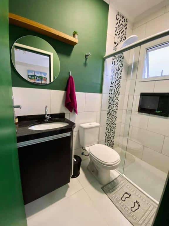 Banheiro do Aparthouse Maranduba com uma pia para lavar mão de mármore preto. um espelho redondo na parede, uma toalha pendurada e ao lado um vaso sanitário. Do lado tem um box de vidro com o chuveiro e uma janela. Imagem ilustrando post Airbnb em Maranduba.