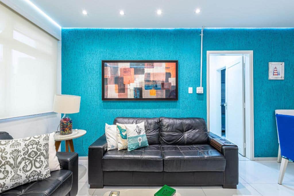 Sala no Apê Descolado no Guarujá – Enseada. As paredes são azuis e brancas. Há um sofá preto de couro no centro, e atrás dele há um quadro e uma porta para outro cômodo. Na esquerda há outro sofá igual e atrás há uma janela.