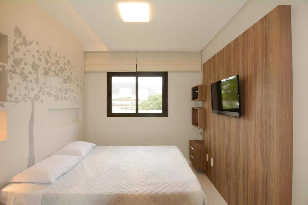 Quarto do airbnb Apt Jurerê. Do lado direito há um painel de madeira com uma televisão presa, já na esquerda observa-se a cama de casal e ao fundo uma janela com persianas. Imagem utilizada para ilustrar o post airbnb em Jurerê Internacional.