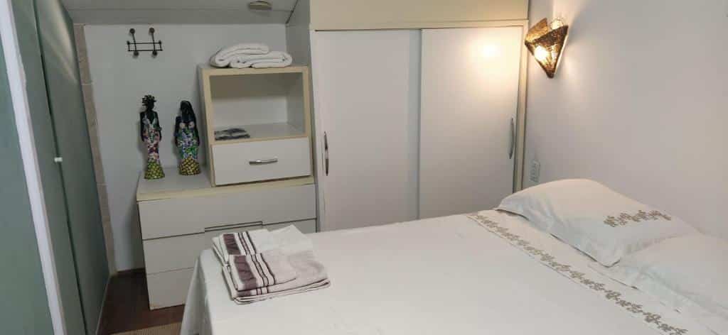 Foto do quarto em Apartamento 1 suíte garagem Praia do Forte 500m da Praia, que ilustra o post de airbnb na Praia do Forte. A cama de casal está no centro e do lado dela há um guarda-roupa com duas portas e algumas gavetas. Tudo no quarto é branco.