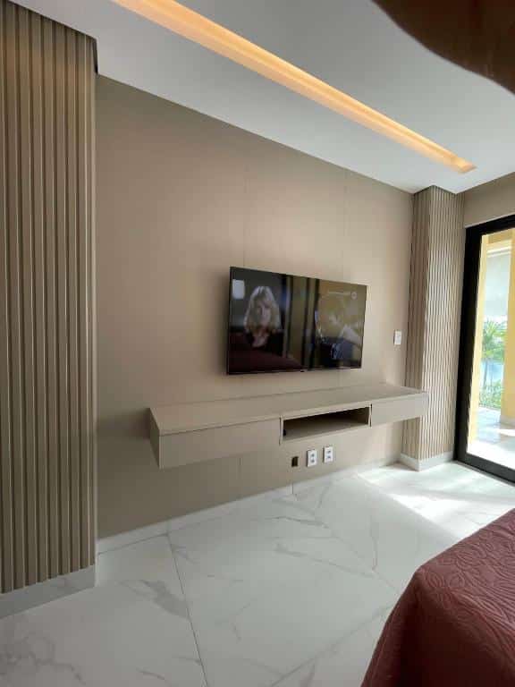 Outro ângulo do quarto no Apartamento Alto Padrão 3 quartos Iberostate. De frente para a acama há uma TV fixa na parede, e na direita há uma porta de vidro com vista para o exterior.