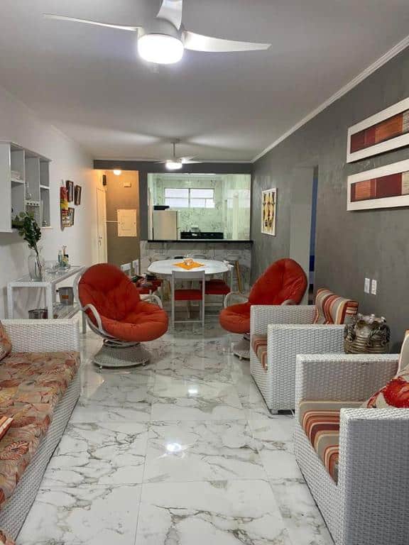 Sala bem decorada em ton de cinza, branco e vermelho no Apartamento Guarujá Pitangueiras. A sala possui um sofá na esquerda e duas poltronas na direita. Entre eles há mais duas poltronas. No fundo há uma área de jantar e a cozinha.
