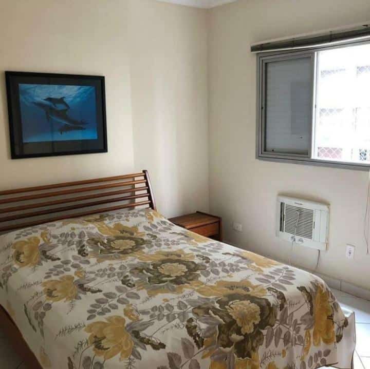 Imagem do quarto do Apartamento na Praia de Pitangueiras-Guarujá, ilustrando o post sobre airbnb em Guarujá. A cama de casal está em foco, e do lado direito dela há uma janela com rede.