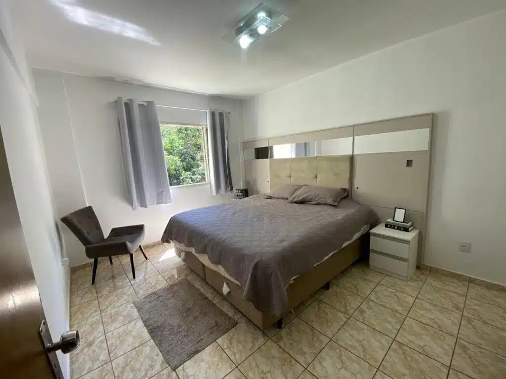 Foto do quarto em Apartamento VIP Centro Serra Negra. O quarto é grande e possui uma cama box de casal com cabeceira espelhada no centro. Na esquerda da cama há uma janela e uma poltrona preta.
